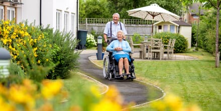 Man Pushes Women in Specialist Wheelchair Along Garden Path.