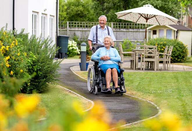 Man Pushes Women in Specialist Wheelchair Along Garden Path.