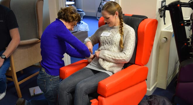 Woman Seated in Red Chair, Whilst Being aided by another. 