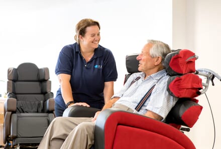 Care Assistant Aids Elderly Man in Red Disability Chair.